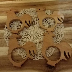 May include: Four wooden rooster-shaped napkin rings. The napkin rings are light brown and sit on a white lace doily. The background is a wooden surface.