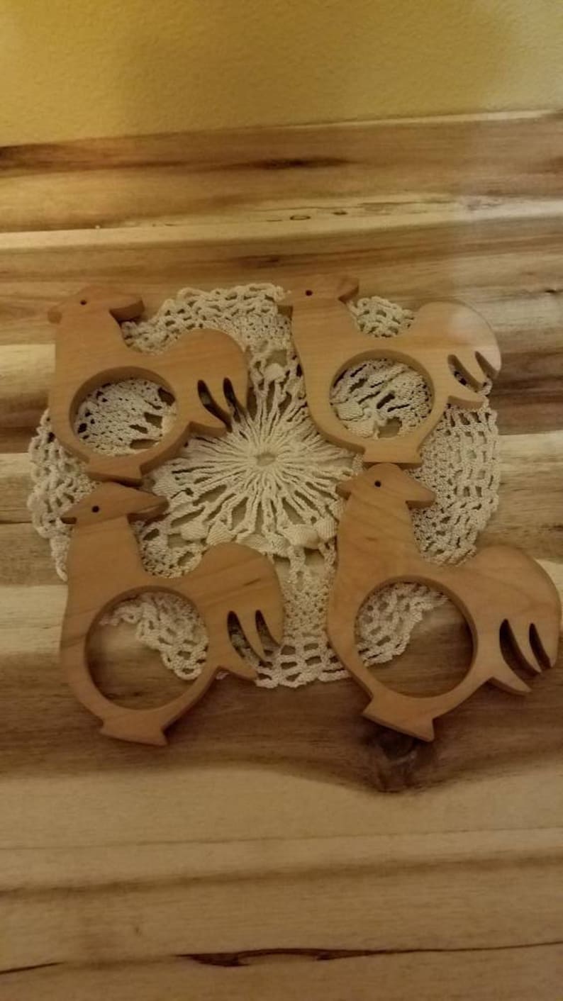 May include: Four wooden rooster-shaped napkin rings. The napkin rings are light brown and sit on a white lace doily. The background is a wooden surface.