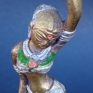 May include: A gold-colored statue of a woman in a green and white patterned dress, with a red dot on her forehead. The statue is in a dancing pose with one arm raised.