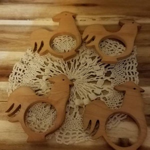 May include: Five wooden rooster-shaped napkin rings arranged on a cream-colored lace doily. The napkin rings are light brown and have a cutout design. The background is a wooden surface.
