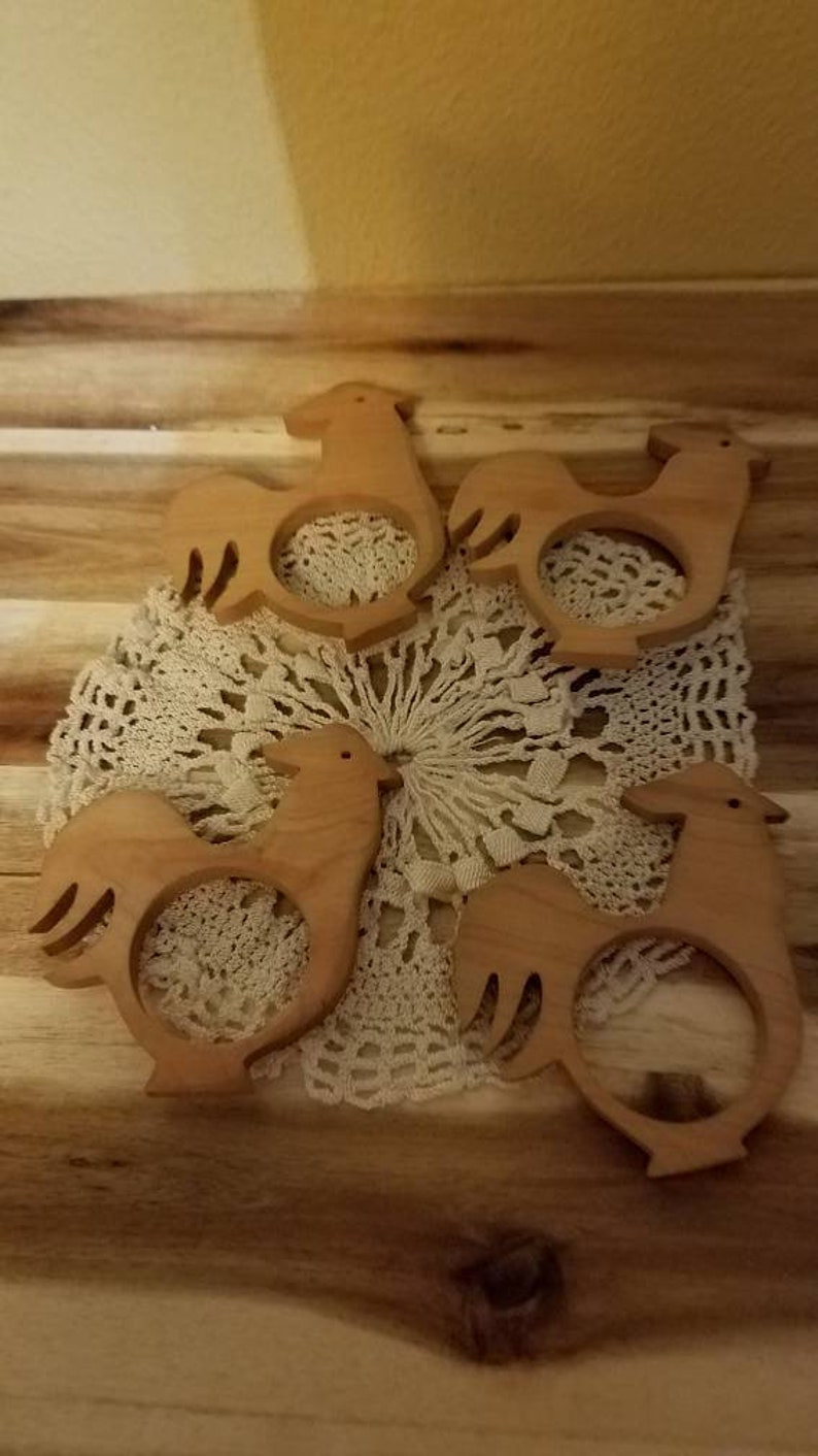 May include: Five wooden rooster-shaped napkin rings arranged on a cream-colored lace doily. The napkin rings are light brown and have a cutout design. The background is a wooden surface.