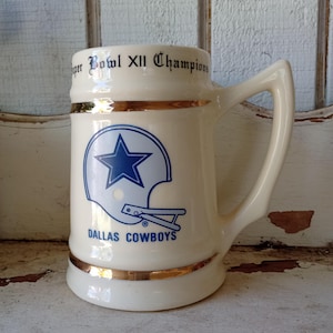 May include: Cream-colored ceramic mug featuring the Dallas Cowboys logo, a blue star inside a white helmet, and the words "DALLAS COWBOYS." The mug has gold trim and the words "Super Bowl XII Champions" around the rim.