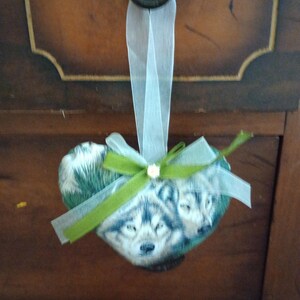 May include: Heart-shaped ornament featuring a wolf design, accented with a green bow and sheer white ribbon. The ornament is suspended from a dark, ornate knob, creating a rustic and decorative appeal.