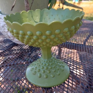 May include: A green hobnail glass pedestal bowl. The bowl has a scalloped rim and is covered in small, rounded bumps. The pedestal base is also covered in the same hobnail pattern. The bowl is sitting on a metal table.