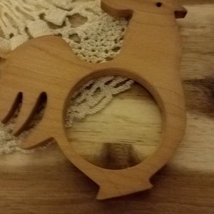 May include: Wooden rooster-shaped napkin ring. The light brown ring features a cutout design and a simple, rustic aesthetic. The rooster has a small eye and a curved tail. The napkin ring is placed on a wooden surface with a lace doily.