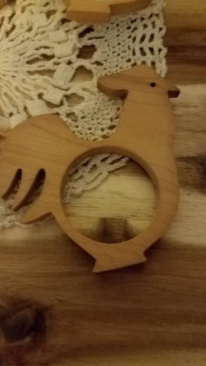 May include: Wooden rooster-shaped napkin ring. The light brown ring features a cutout design and a simple, rustic aesthetic. The rooster has a small eye and a curved tail. The napkin ring is placed on a wooden surface with a lace doily.