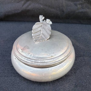 May include: A silver trinket box with a leaf-shaped lid. The lid is detailed with a raised leaf design and a small stem.