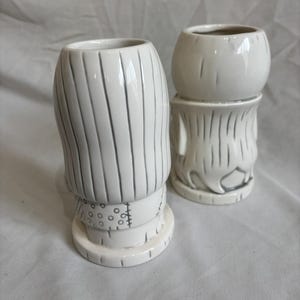 May include: Two white ceramic tiki mugs. One is tall with vertical lines and a stitched band. The other is shorter, with a round top and a similar design. Both mugs have a glossy finish and are on a white surface.
