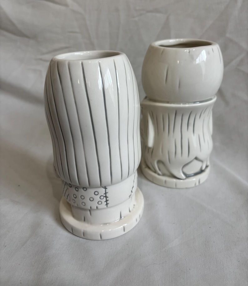 May include: Two white ceramic tiki mugs. One is tall with vertical lines and a stitched band. The other is shorter, with a round top and a similar design. Both mugs have a glossy finish and are on a white surface.