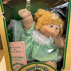 May include: A Cabbage Patch Kids doll in its original box. The doll has blonde yarn hair, blue eyes, and a light green dress with white lace trim. The box includes a birth certificate and adoption papers.