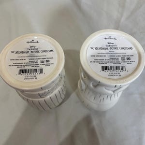 May include: Two white ceramic mugs with black vertical lines and facial features, from Tim Burton's The Nightmare Before Christmas. The mugs have a circular label on the bottom with the Hallmark and Disney logos.