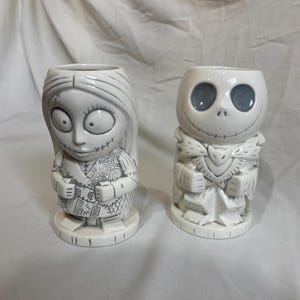 May include: Two white ceramic character mugs. One mug is Sally from The Nightmare Before Christmas, with stitched details. The other is Jack Skellington, with a skeletal face and a bat-shaped bow tie. Both mugs have a cylindrical shape.