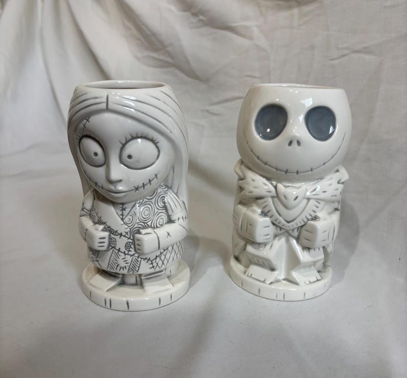 May include: Two white ceramic character mugs. One mug is Sally from The Nightmare Before Christmas, with stitched details. The other is Jack Skellington, with a skeletal face and a bat-shaped bow tie. Both mugs have a cylindrical shape.