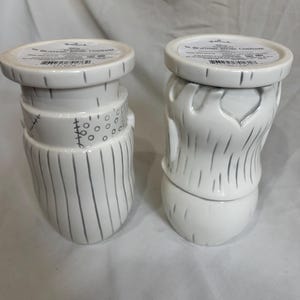 May include: Two white ceramic candle holders, inspired by "The Nightmare Before Christmas." One has a stitched neck and vertical lines, the other a beard design. Both have a round top with text. The holders are decorative.