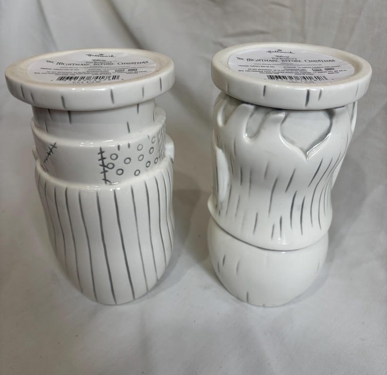 May include: Two white ceramic candle holders, inspired by "The Nightmare Before Christmas." One has a stitched neck and vertical lines, the other a beard design. Both have a round top with text. The holders are decorative.