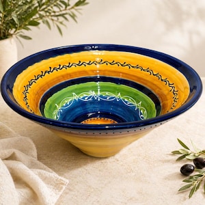 May include: A large, hand-painted ceramic bowl with a vibrant, concentric design. The bowl features bands of yellow, orange, green, and blue, with decorative black and white accents. The bowl's exterior is a pale yellow.