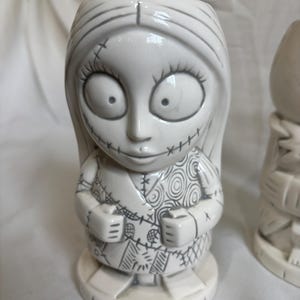 May include: A white ceramic tiki mug in the shape of a character with large eyes, stitched features, and long hair. The mug has a detailed design with grey outlines and stands on a round base. A unique piece of drinkware.