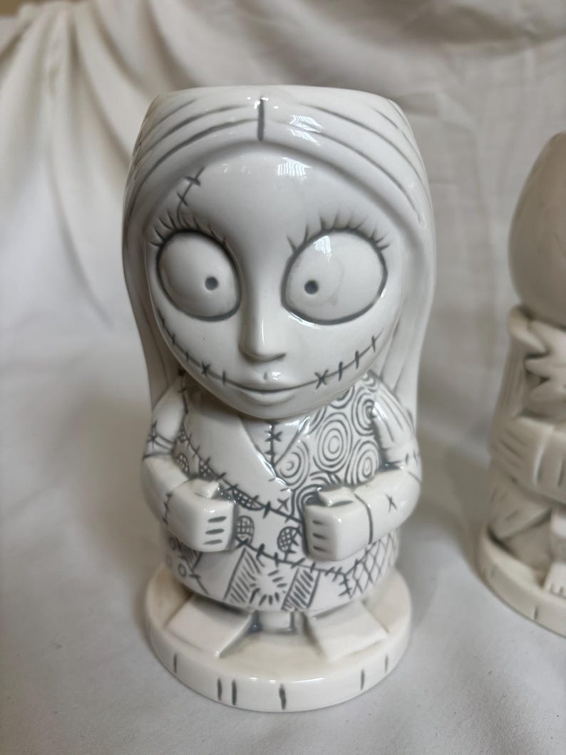 May include: A white ceramic tiki mug in the shape of a character with large eyes, stitched features, and long hair. The mug has a detailed design with grey outlines and stands on a round base. A unique piece of drinkware.