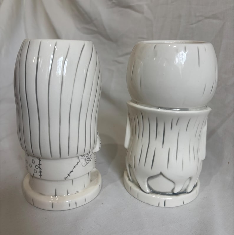 May include: Two white ceramic mugs with a Frankenstein's monster design. The mugs have a textured surface with vertical lines and grey accents. One mug is taller than the other, and both have a round base.