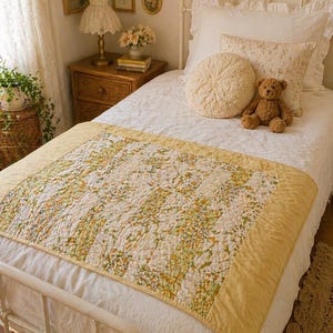 May include: A cozy bedroom scene featuring a white bed with a floral quilt and pillows. A small teddy bear sits on the bed. The quilt has a yellow border and a pattern of green, yellow, and white floral designs. A nightstand with a lamp and framed artwork complete the scene.