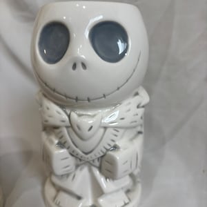May include: White ceramic tiki mug depicting Jack Skellington's face with grey eyes and stitched mouth. The mug has a carved design on the body, with a stylized collar and a base. A unique collectible for fans of the film.