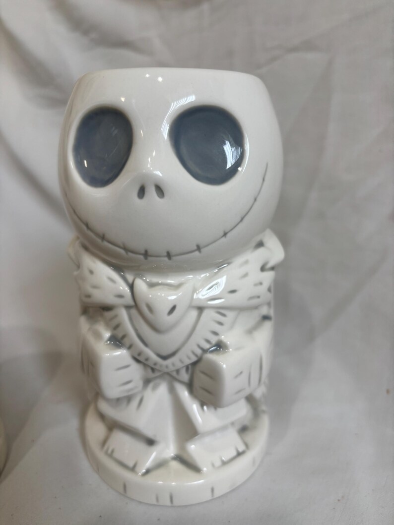 May include: White ceramic tiki mug depicting Jack Skellington's face with grey eyes and stitched mouth. The mug has a carved design on the body, with a stylized collar and a base. A unique collectible for fans of the film.