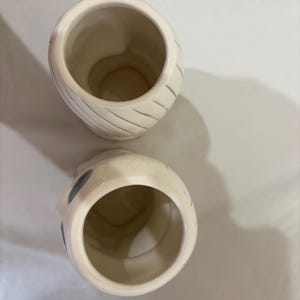 May include: Two off-white ceramic mugs with a mummy-like design. Each mug has a wide opening and a rounded body. Gray diagonal lines wrap around the body of one mug. The other mug has gray circles on the side.