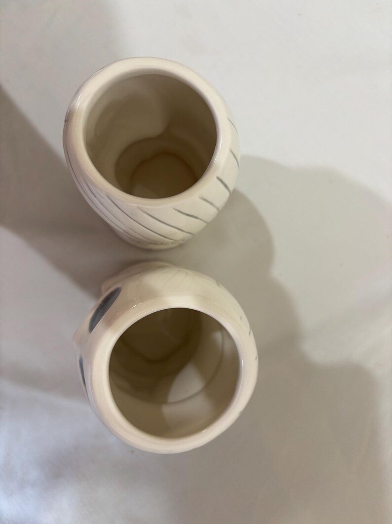 May include: Two off-white ceramic mugs with a mummy-like design. Each mug has a wide opening and a rounded body. Gray diagonal lines wrap around the body of one mug. The other mug has gray circles on the side.