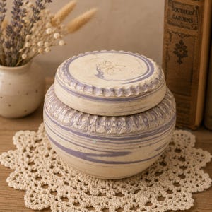 May include: A round ceramic jar with a lid, featuring a marbled pattern of cream and muted purple swirls. The jar has a textured, scalloped rim detail around the lid and the upper body, with a small embossed floral design on the center of the lid.