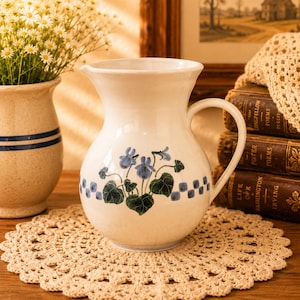 May include: A cream-coloured ceramic jug with a handle, adorned with blue floral and green leaf patterns. A blue and white chequered design borders the floral pattern. The jug sits on a beige doily, with a vase of flowers and books in the background.