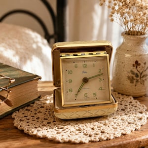 May include: A square, gold-toned travel alarm clock with a cream-colored, quilted-texture case. The clock face features green Arabic numerals, the brand name Artec, and the word Luminous, with green hands indicating the time on a white background.