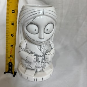 May include: A white ceramic Sally figure from The Nightmare Before Christmas. The figure is approximately 15 cm tall and features black stitch marks and details. The figure is standing on a white base.