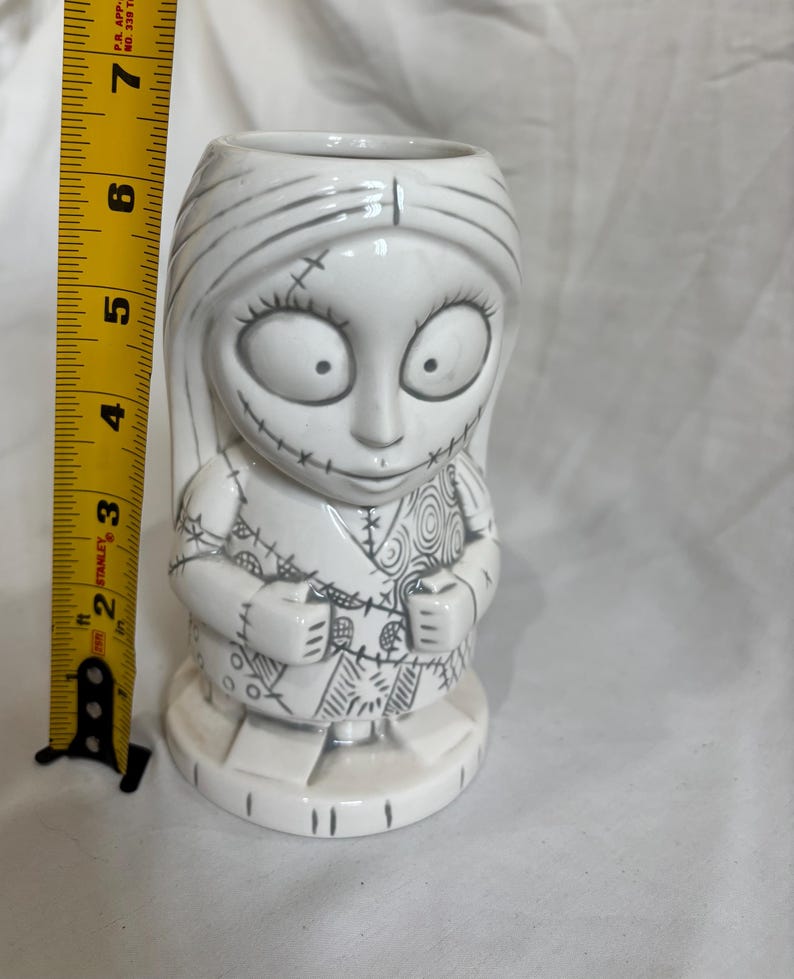May include: A white ceramic Sally figure from The Nightmare Before Christmas. The figure is approximately 15 cm tall and features black stitch marks and details. The figure is standing on a white base.