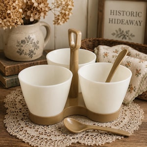 May include: A set of three white ceramic bowls held in a tan plastic stand with a central handle. The stand features a base that supports the bowls, and the set includes two matching tan plastic spoons for serving, arranged on a round lace doily.