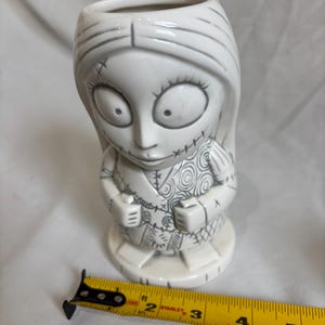 May include: A white ceramic mug shaped like Sally from The Nightmare Before Christmas. The mug features Sally's stitched design and facial features, with grey detailing. The mug is approximately 13 cm tall.