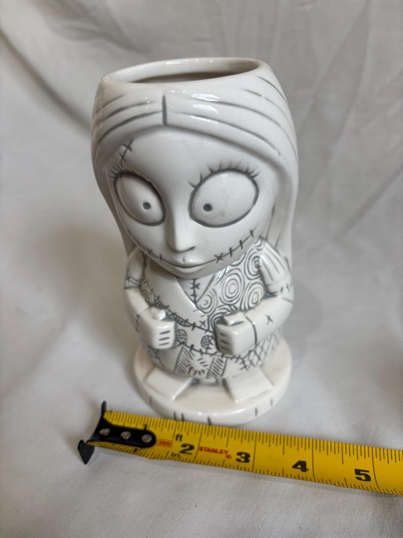May include: A white ceramic mug shaped like Sally from The Nightmare Before Christmas. The mug features Sally's stitched design and facial features, with grey detailing. The mug is approximately 13 cm tall.