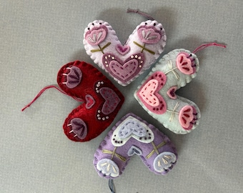 May include: Four felt heart ornaments with embroidered floral designs. The hearts are in shades of purple, pink, and red.