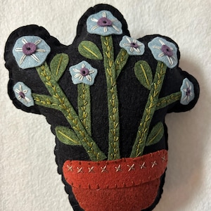 May include: A felt flower pot with blue flowers and green leaves. The pot is red and has a black background.
