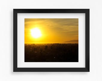 May include: Framed print of a vibrant sunset. The sun is a bright, golden orb in a yellow and orange sky. Silhouetted trees and a mountain range are visible in the distance, creating a scenic landscape.