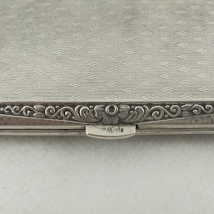 Op de afbeelding: Close-up van een zilveren sigarettenetui. De behuizing heeft een getextureerd oppervlak met een herhalend patroon. De randen zijn versierd met een sierlijk bloemen- en scrollontwerp. De sluiting is zichtbaar met de initialen "WM" en "@obe".