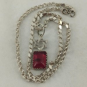 May include: A silver necklace featuring a Star of David pendant and a rectangular red gemstone. The chain is a delicate, textured design. The pendant is suspended from a small ring. The necklace is displayed on a white surface.