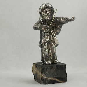 Może przedstawiać: Srebrna figurka mężczyzny grającego na skrzypcach. Figurka stoi na ciemnej, prostokątnej kamiennej podstawie. Mężczyzna ma na sobie kapelusz i płaszcz, a smyczek skrzypiec jest złoty. Figurka ma około 10 cm wysokości.