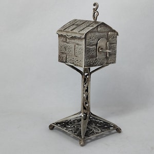 May include: A silver-toned metal miniature house sculpture featuring a textured brick-patterned exterior, a small hinged door with a latch, and a decorative chimney. The structure is supported by a tall, ornate pedestal base with openwork.