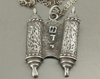 May include: A silver-toned pendant necklace featuring a stylized Torah scroll design. The pendant has two scroll-shaped elements with textured surfaces and a central panel with Hebrew letters. The necklace chain is also silver-toned.