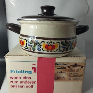 Pode incluir: Uma panela de cozinha vintage bege e castanha com tampa e alças. A panela apresenta uma faixa floral decorativa em vermelho, amarelo e azul. Está sobre uma caixa com texto em alemão.