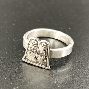 May include: A silver ring featuring a detailed depiction of the Ten Commandments tablets. The ring has a polished band and the tablets are engraved with Hebrew text. The ring is likely made of sterling silver.