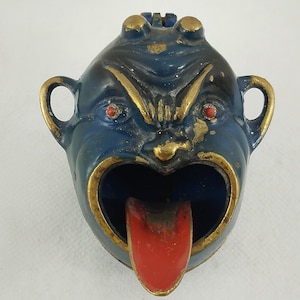 Puede incluir: Un cenicero vintage con forma de cara de demonio azul con detalles dorados. El cenicero tiene ojos rojos y una lengua roja. Tiene dos pequeñas asas a los lados.