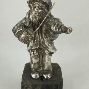 May include: A silver-colored figurine of a man playing a violin. The figure is wearing a hat and coat, and is standing on a dark, textured base. The violin bow is a contrasting gold color.