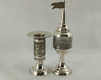 May include: Two silver-toned decorative items. One is a candlestick with an openwork design and Hebrew text. The other is a tall, ornate piece with a flag on top. Both items have a shiny, reflective surface and are likely made of metal.