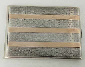 Op de afbeelding: Een rechthoekige zilveren sigarettenetui met een decoratief patroon. De etui heeft afwisselende banden van zilver en goudkleurig metaal. De zilveren banden hebben een gestructureerd, herhalend ontwerp, terwijl de gouden banden glad zijn.